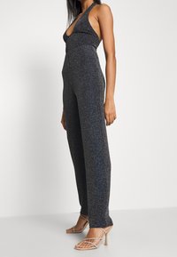 Glittrig svart jumpsuit med halterneck, djup V-halsringning och vida ben. Strukturerad tyg fångar ljuset. Matchad med klara klacksandaler.