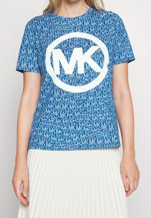 Blauw shirt met korte mouwen met herhaalde witte "MK" letters en een grote witte "MK" logo in een cirkel, gedragen met een witte geplooide rok.