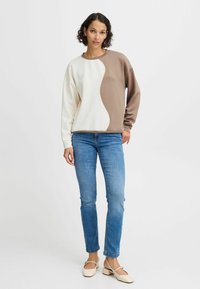 Sweatshirt à col rond en coton crème et marron, avec un design fluide et une texture douce. Porté avec un jean bleu et des chaussures claires.