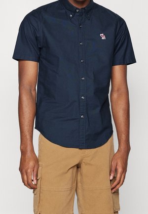 Hombre vestido con una camisa azul marino de manga corta con botones y pantalones cortos cargo color beige, de pie con los brazos relajados a los lados.