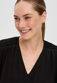Zwarte blouse met een V-hals, geplooide textuur op de schouders en een gladde stof. Het heeft een relaxte pasvorm die geschikt is voor casual gebruik.