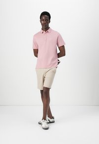 Ροζ πόλο μπλούζα με γιακά και μικρό λογότυπο, συνδυασμένη με μπεζ σορτσάκι και γκρι αθλητικά παπούτσια, σε λευκό φόντο.