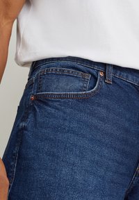 Mörkblå denimjeans med en åtsittande design, som har en bakficka och koppardetaljer. Tyget har en slät textur och en lätt stretch.