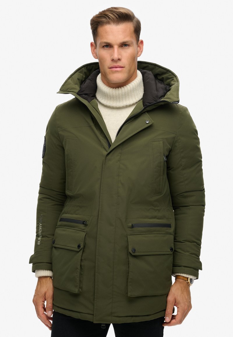 Moški v olivno zeleni zimski jakni s kapuco, oblečen v kremasto pulover s turtleneck ovratnikom, stoji pred enobarvnim belim ozadjem.