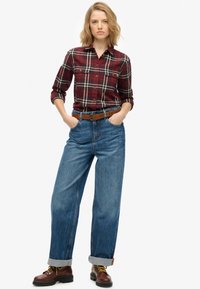 Camisa de franela roja con un patrón de cuadros en blanco y negro, combinada con jeans de pierna ancha azul y botas marrones con cordones.