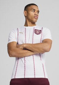 Camisa desportiva branca com riscas verticais em encarnado, apresentando um pescoço redondo, mangas curtas, logótipo dourado da Puma e um emblema do clube em encarnado no peito.