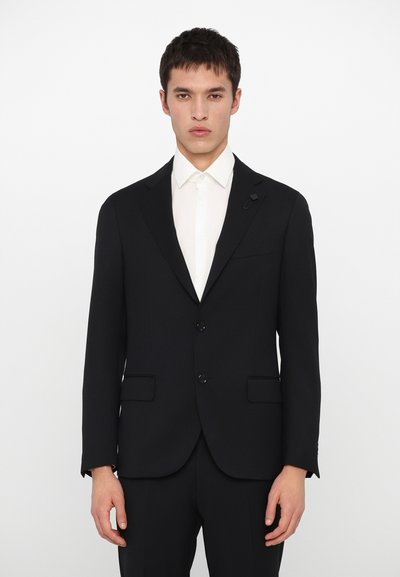 Chaqueta de traje negra a medida con solapas de muesca, dos bolsillos delanteros y una silueta elegante. Lleva puesta una camisa de vestir blanca.