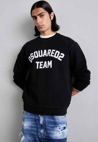 Swearshirt negru cu textul "DSQUARED2 TEAM" în litere albe, îndrăznețe. Puloverul are un guler rotund și manșete ribbate, asociat cu blugi albaștri ușor distruși.