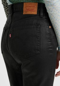 Svarta denimjeans med hög midja, med en läderpatch på ryggen, arcuat sömnad på fickorna och en röd etikettlogotyp. Texturerad yta.