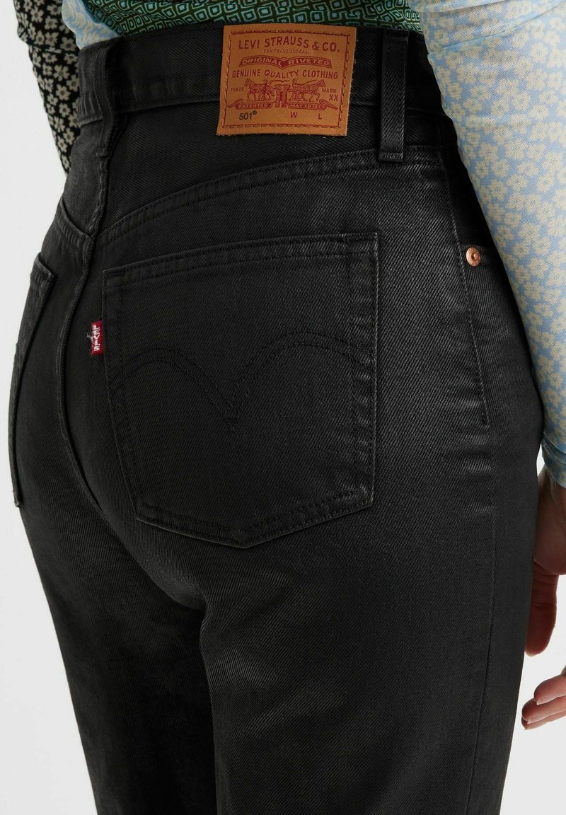 Jean en denim noir à taille haute, avec un patch en cuir au dos, des coutures en arc sur les poches et un logo en forme de tab rouge. Surface texturée.