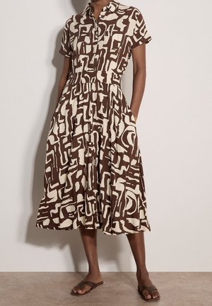 Femme portant une robe chemise marron et beige à motif abstrait, manches courtes et poches, associée à des sandales plates marron.