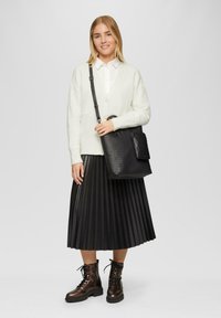 Weiße Strickjacke, vorne geknöpft, kombiniert mit einem schwarzen Faltenrock. Als Accessoires dienen eine strukturierte schwarze Tasche und braune Schnürstiefel.
