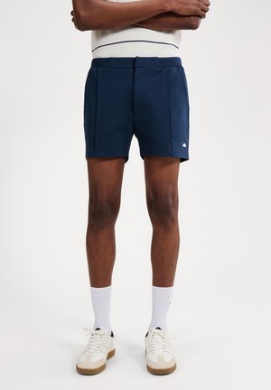 Man met marineblauwe korte broek, witte sportsokken en witte sneakers, met gekruiste armen staand voor een effen achtergrond.