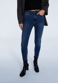 Jeans ajustados de color azul oscuro con talle alto, que presentan un diseño clásico de cinco bolsillos y costuras sutiles; combinados con botas de tacón alto negras.