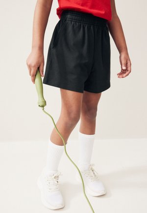 Dziecko ubrane w czarne spodenki, białe skarpetki i białe trampki trzymające zieloną rączkę skakanki, z czerwoną koszulką widoczną w talii.