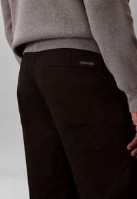 Detail osoby v tmavých nohaviciach s etikietou Calvin Klein a sivom pletenom svetri, zobrazená zozadu s uvoľnenou rukou pozdĺž tela.