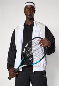 Tennisoutfit bestående af en sort og hvid lynlåsjakke, sort T-shirt og sorte bukser. Holder en tennisracket med blå detaljer.