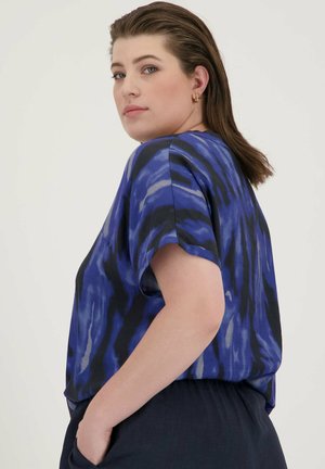 Blouse ample avec un motif abstrait bleu et noir, manches courtes, tissu léger, poche latérale visible sur un pantalon foncé.