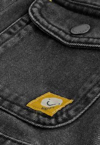 Tissu en denim noir en gros plan avec une poche à rabat, un bouton noir et un petit patch jaune avec un visage souriant blanc et le mot "Happy."