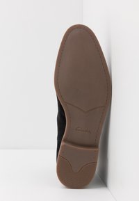 Černé semišové boty s hladkou hnědou gumovou podrážkou. Mají zaoblenou špičku, mírně texturované polstrování a nenápadné logo Clarks na podrážce.