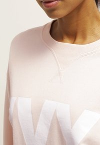 Sudadera de color rosa claro con cuello redondo y textura acanalada. Presenta un gran texto blanco en el frente, mostrando las letras "NY."