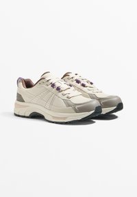 Béžové sportovní boty s texturovanou síťovinou a syntetickými překrytími. Mají fialové akcenty, kulaté tkaničky a polstrovanou podrážku.