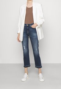 Blazer bianco sopra un top marrone, abbinato a jeans blu scuro strappati con orli frangiati e sneaker bianche.