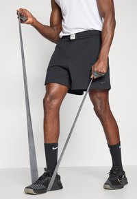 Μαύρες αθλητικές βερμούδες με ελαστική ζώνη, συνδυασμένες με μαύρες κάλτσες και παπούτσια Nike. Μια γκρί ταινία αντίστασης κρατείται με το ένα χέρι.