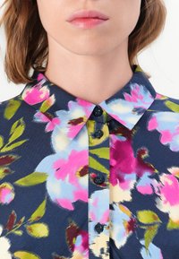 Woman wearing a navy blue shirt with a floral pattern in pink, blue, green, and cream, featuring a pointed collar and black buttons.