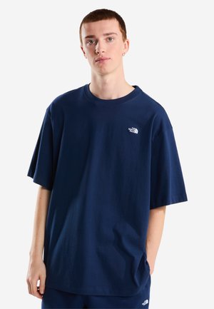 Junger Mann, der ein lockeres dunkelblaues The North Face T-Shirt und passende Hose trägt, steht mit einer Hand in der Tasche vor weißem Hintergrund.