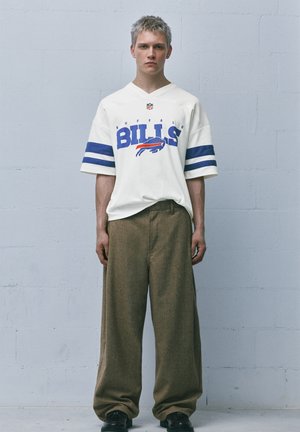 Giovane uomo con capelli corti che indossa una maglia bianca dei Buffalo Bills con strisce blu e pantaloni marroni larghi, posato contro un muro semplice.