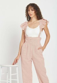 Mujer con cabello rizado, vestida con una blusa blanca y pantalones de talle alto a rayas rosas con tirantes, de pie junto a un taburete blanco sobre un fondo liso.