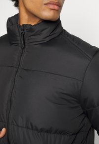 Giacca a vento nera imbottita con colletto alto, chiusura con zip frontale e texture trapuntata. Presenta una finitura liscia e lucida e dettagli di cucitura orizzontale.