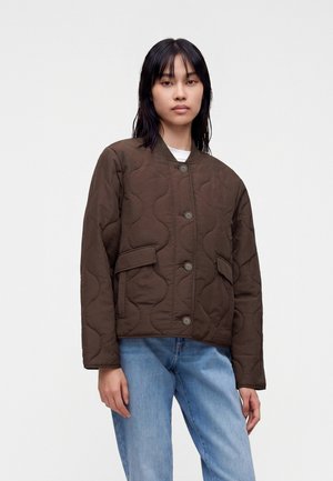 Jeune femme portant une veste matelassée marron foncé avec des boutons et un jean bleu clair, debout devant un fond uni.
