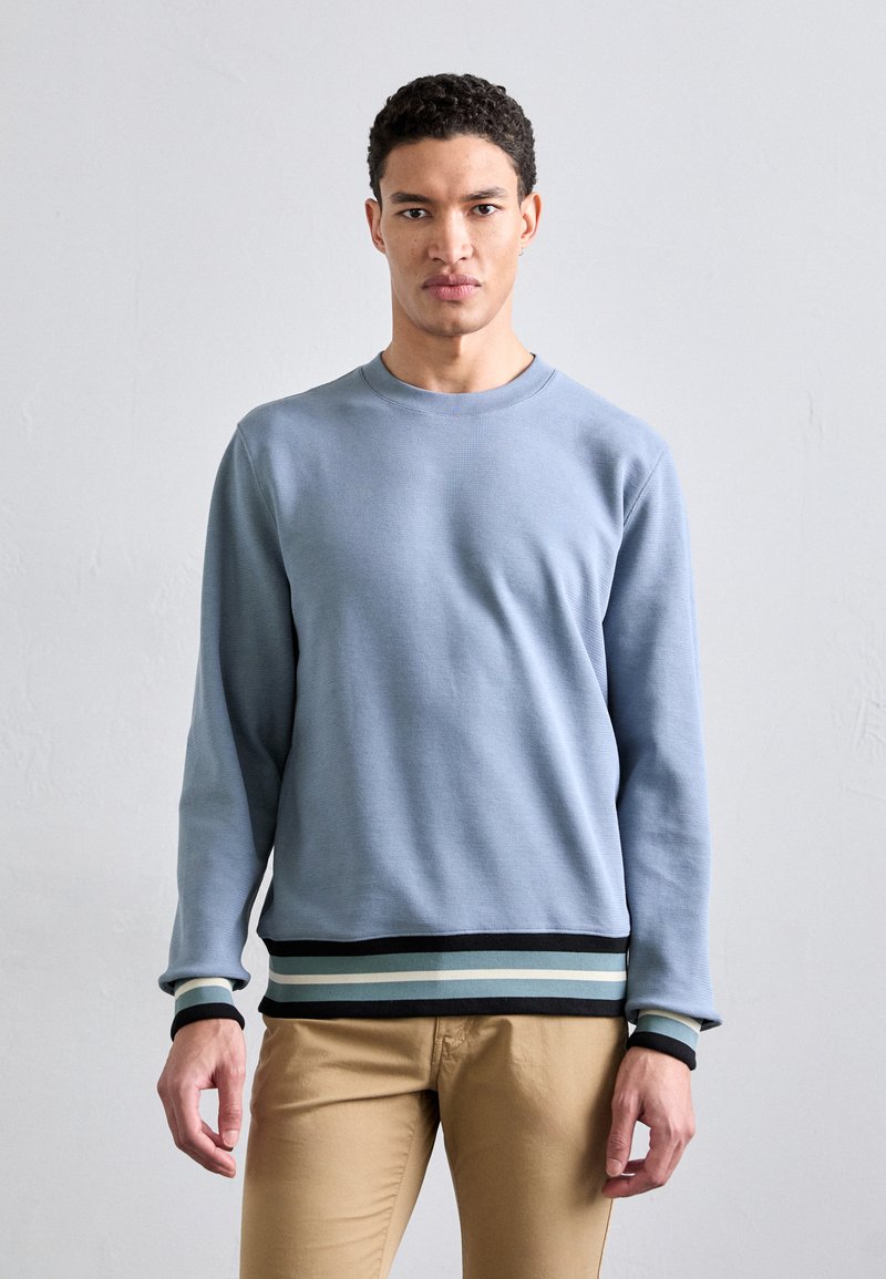 Jasnoniebieski sweter crewneck z ribbowanymi mankietami i dołem, z czarnymi oraz dwukolorowymi paskami na dole. Miękka tekstura i luźny krój.