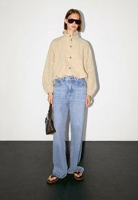 Beżowa lniana koszula z wysokim kołnierzem i zapięciem na guziki, zestawiona z jasnoniebieskimi szerokimi spodniami dżinsowymi i ciemnobrązowymi klapkami. Do zestawu dołączona jest brązowa torebka.