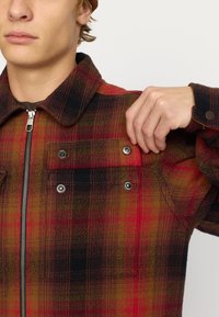 Rood en zwart geruite jas met een ritssluiting aan de voorkant, drukknoopzakken en een kraag. Zacht, gestructureerd materiaal met contrasterende kleuren.