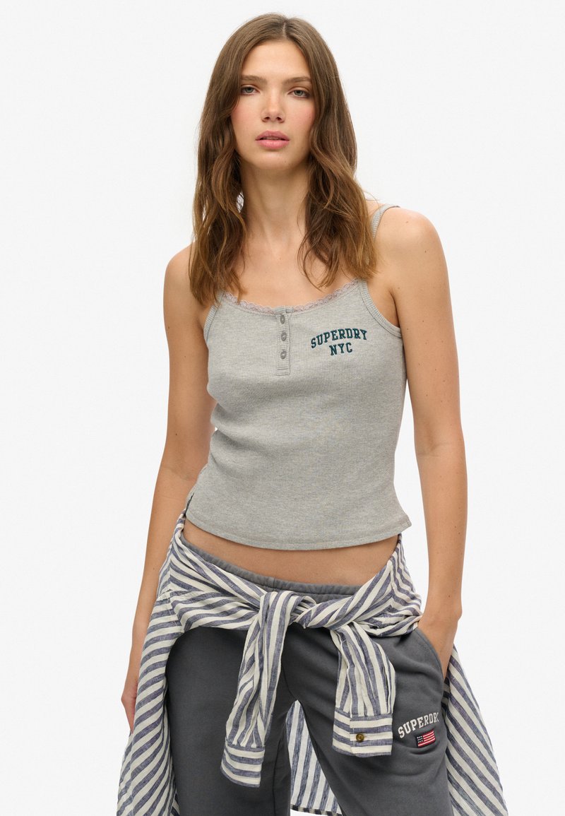 Woman wearing a grey ribbed tank top with "Superdry NYC" text, dark joggers, and a striped shirt tied around the waist, standing with a relaxed pose.