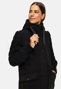 Black coat with a hood, featuring solid wool body and textured knitted sleeves. Includes front pockets and black hardware details.