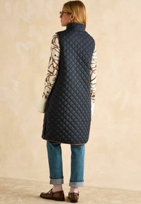 Kvadru gudrs tumši zils vests ar augstu apkakli, ar dimantu rakstu un brūnu apdari, uzvilkts virs rakstainas garām piedurknēm augšējā un ziliem džinsiem.