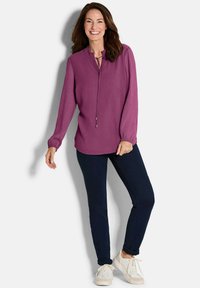 Roze blouse met lange mouwen, een ruchesnek en strikdetail, gecombineerd met donkerblauwe spijkerbroek en lichtgekleurde sneakers.