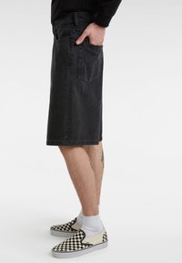 Pantalones cortos de mezclilla negra con corte recto, bolsillos frontales y traseros, y dobladillo sin terminar. Llevados con zapatos slip-on de cuadros.