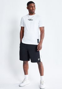 Camiseta blanca con estampado negro "ELEVEN DEGREES", combinada con pantalones cortos negros con franjas laterales blancas y un logo, y zapatillas blancas.