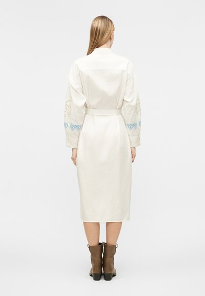 Γυναίκα με ίσια ξανθιά μαλλιά που φοράει ένα κρεμ midi φόρεμα με μπλε κεντημένα μανίκια και καφέ μποτάκια αστραγάλου, στέκεται γυρισμένη με την πλάτη.