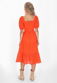 Rochie midi din bumbac roșu, cu mâneci bufante, decolteu pătrat și tiv în straturi. Prezintă detalii fine cu găuri pe parcursul materialului. Purată cu cizme bej.
