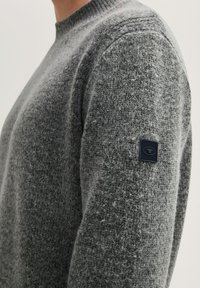 Grauer Strickpullover mit runden Ausschnitt, strukturierter Oberfläche und einem schwarzen Logopatch an der linken Schulter. Warmes und lässiges Design.