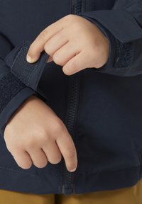 Námořnická bunda s předním zipem a uzavřením na suchý zip na manžetách. Materiál vypadá hladce a odolně, s těsným střihem kolem zápěstí.