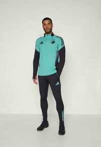 Vīrietis stāv, ģērbies tirkīza un melnā Adidas sporta apģērba komplektā ar augstu apkakli un logotipu uz krūtīm, kas savienots ar melnām Adidas treniņbiksēm un apaviem.