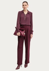 Donna che indossa una blusa bordeaux a maniche lunghe, pantaloni bordeaux a righe, stivali a punta con tacco alto, mentre tiene in mano una borsa a tracolla bordeaux abbinata.