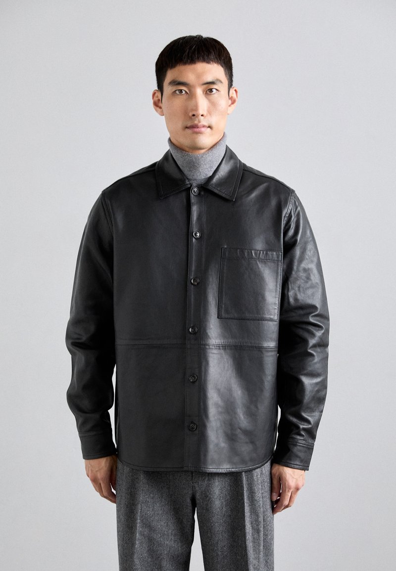 Svart läderjacka med knäppning fram, kontrasterande textur, bröstficka och klassisk krage, matchad med grå byxor.